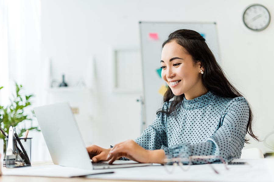 Blog - Young Woman in Polka Dot Blouse Smiles and Uses Her Laptop in a Bright White Office