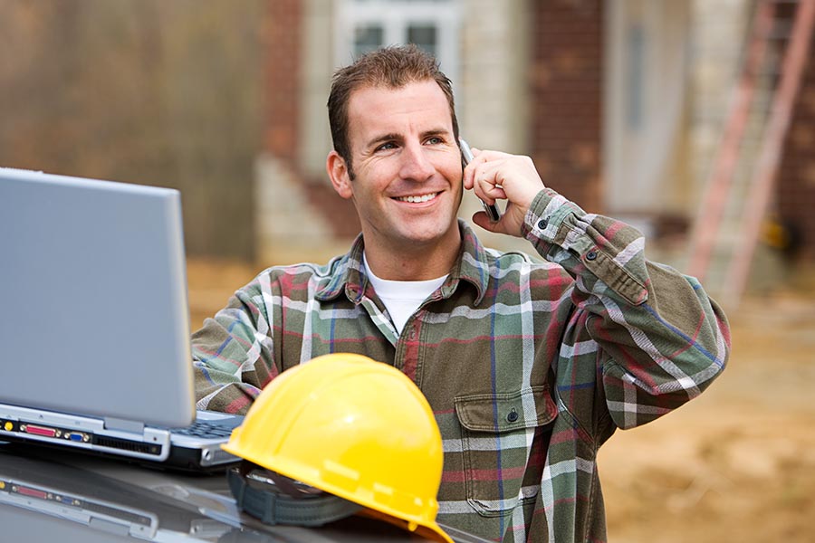 Specialized Business Insurance - Smiling Contractor Makes a Call With His Laptop and Hardhat Propped on Truck Hood, a Residential Job Site Behind Him